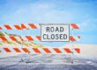 Orange and white barrier with a ROAD CLOSED sign blocks a tarmacked road under a blue sky.